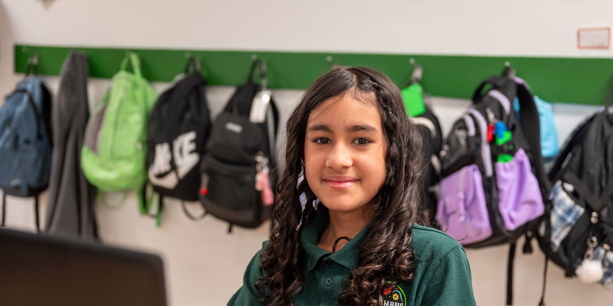Smiling student in a green school polo in front of backpacks on classroom hooks.