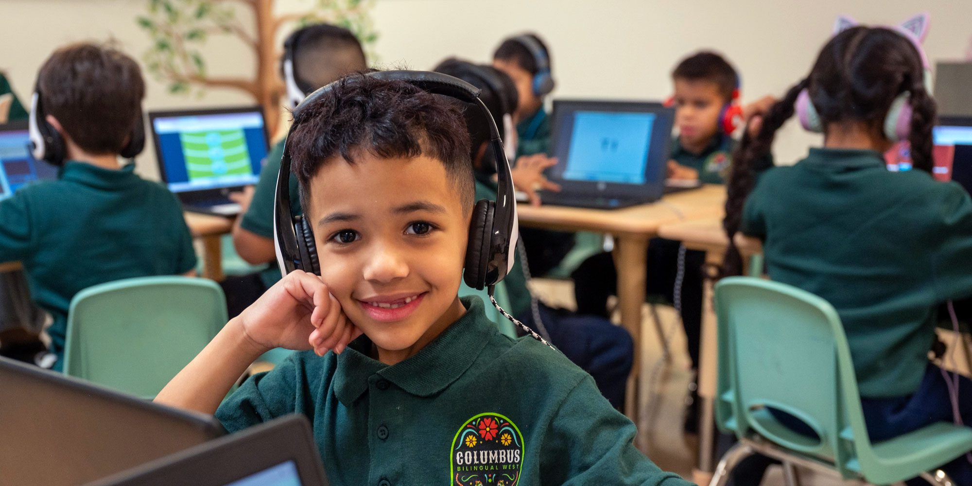Smiling student with headphones in a classroom of students working on laptops.