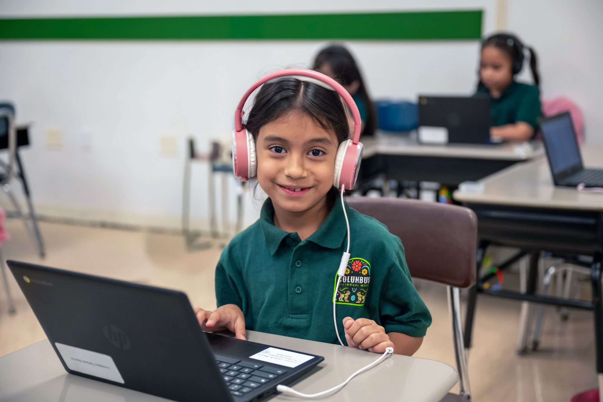 Student wearing pink headphones smiles while working on a laptop.