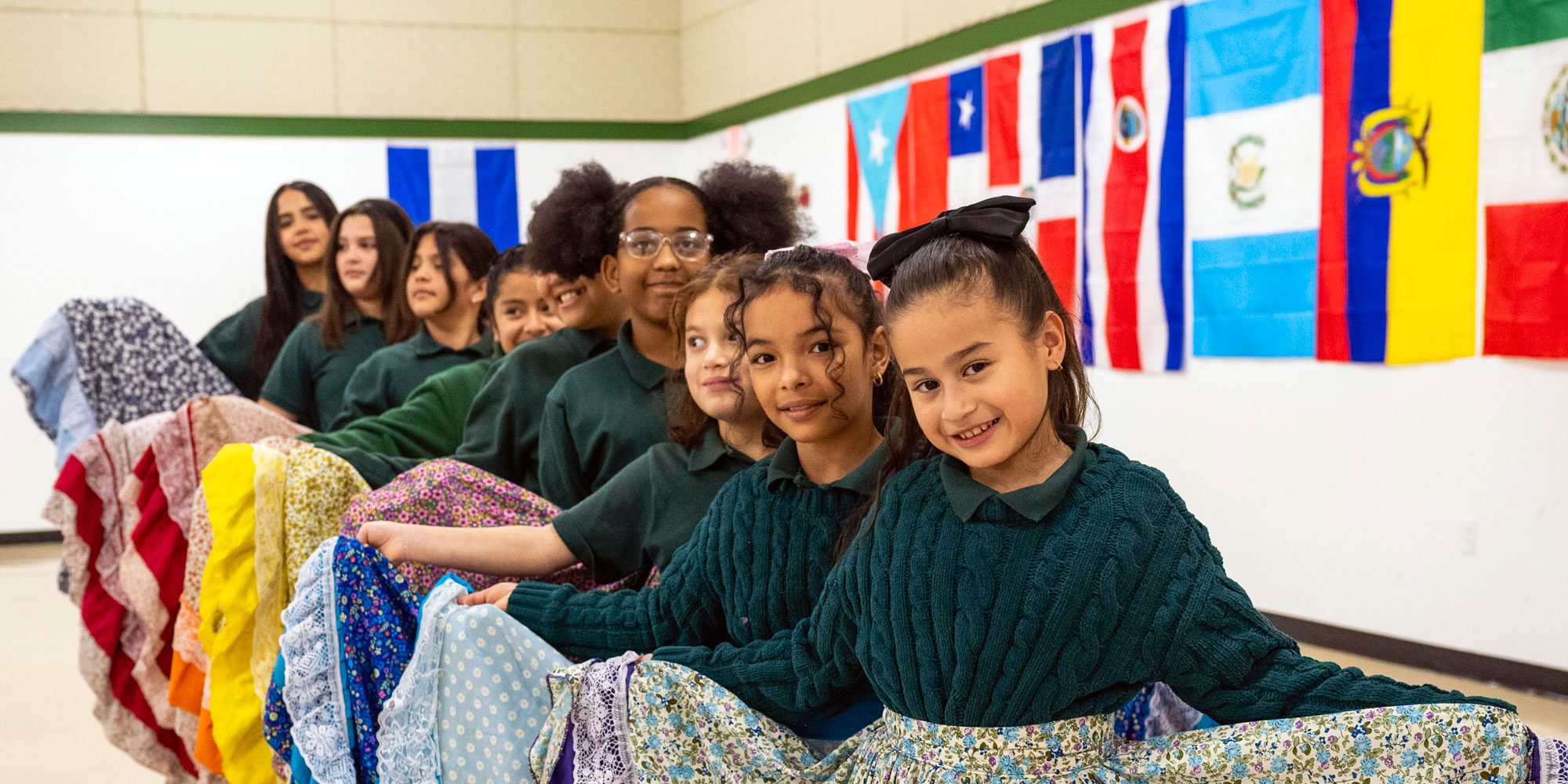 Students in green polos and traditional folkloric skirts seated beneath flags of Latin American countries.
