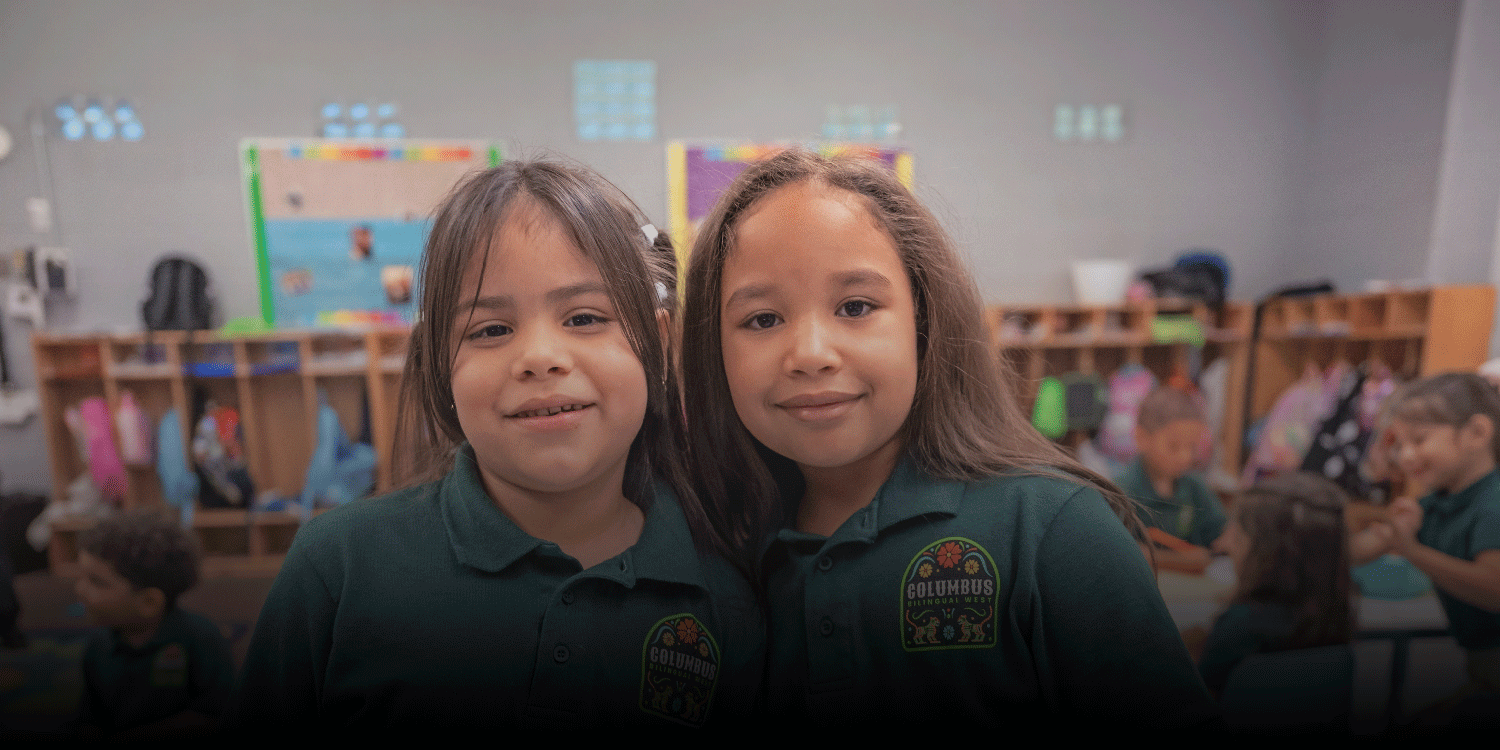 Two smiling students in green school polos in a classroom.